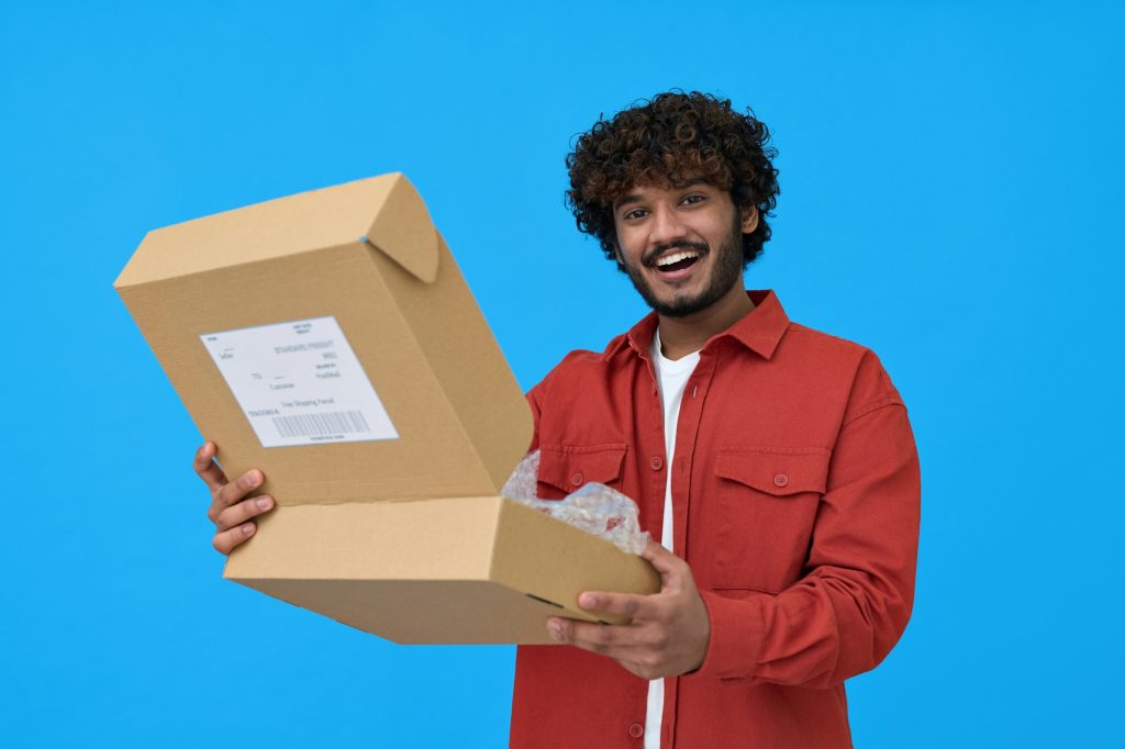 Happy indian young man holding open parcel box isolated on blue background.