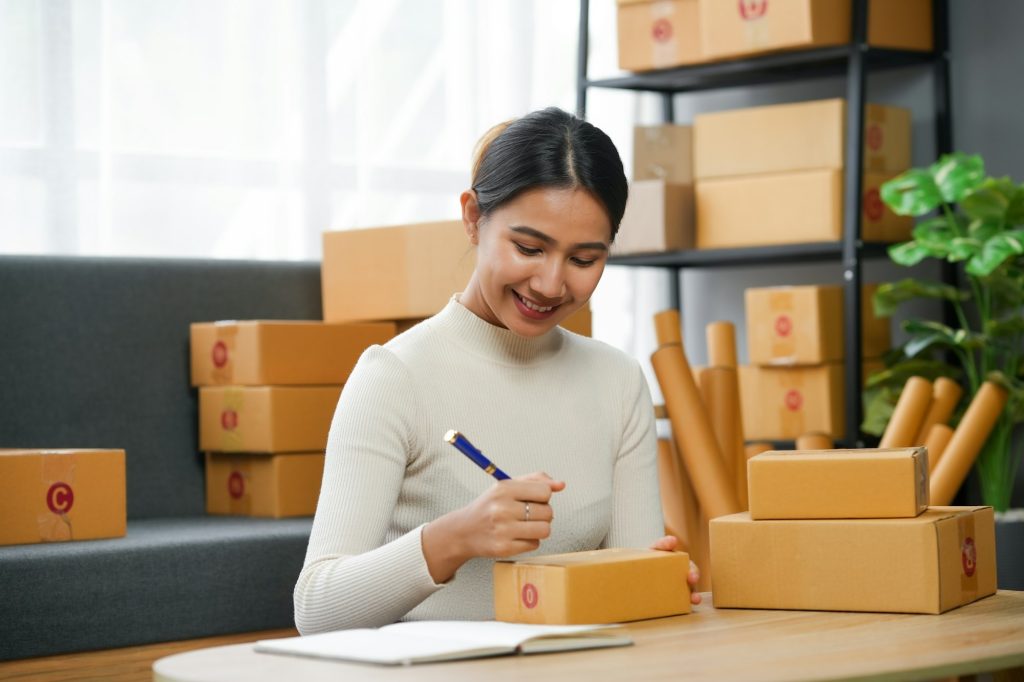 Woman preparing packages for delivery, writing addresses on boxes at home office.