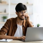 Cheerful indian guy businessman with laptop having phone conversation