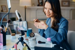 Happy Asian beauty blogger recording makeup tutorial at home.