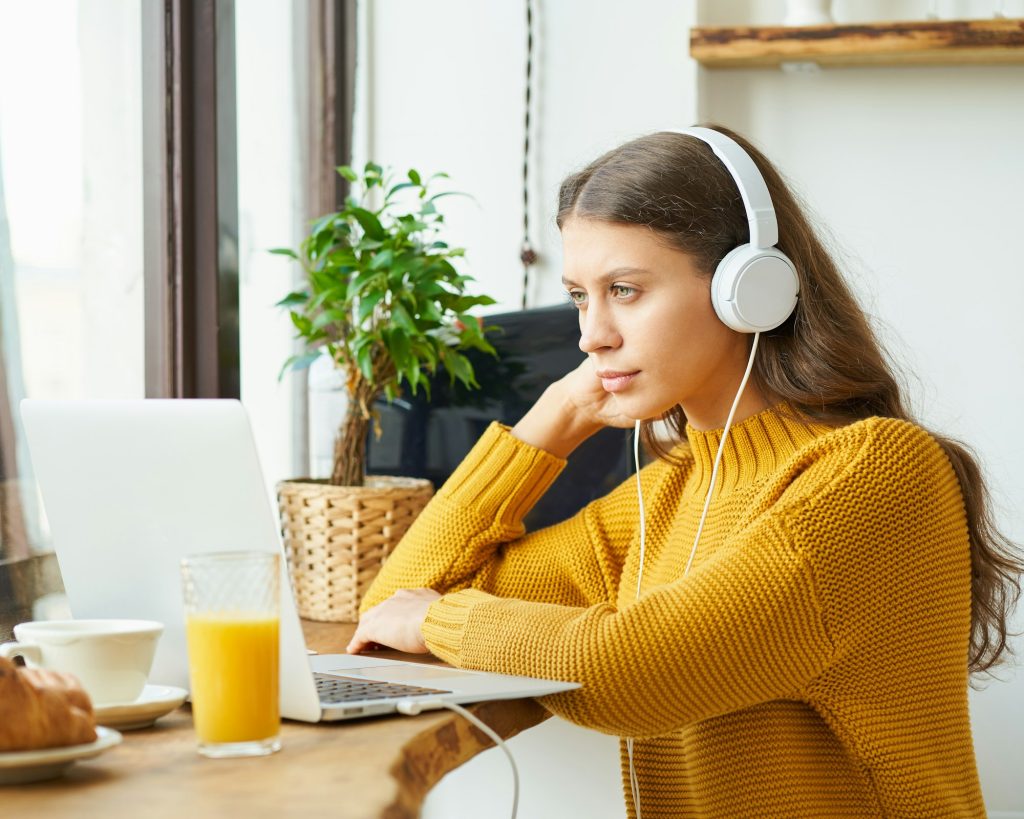 Happy girl in wireless headphones studying online, using laptop and taking breakfast
