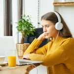 Happy girl in wireless headphones studying online, using laptop and taking breakfast