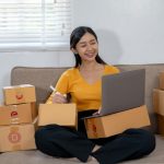Woman working on a laptop and holding a pen, surrounded by packages.