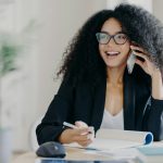 Photo of glad smiling dark skinned woman calls business partner, being in good mood