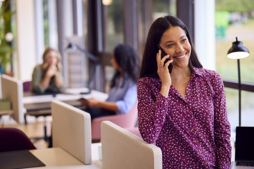 Businesswoman Standing By Partition In Open Plan Office Making Call On Mobile Phone