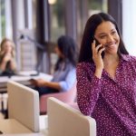 Businesswoman Standing By Partition In Open Plan Office Making Call On Mobile Phone