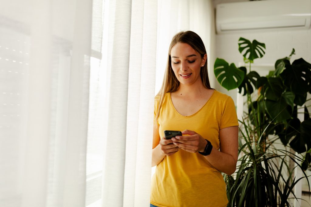 Working woman standing beside a window holding mobile phone. Woman entrepreneur taking a break.