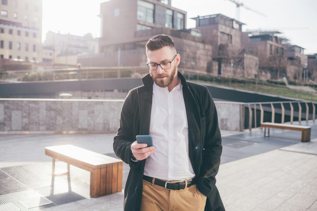 Young bearded man outdoor using smartphone texting searching online buying ecommerce